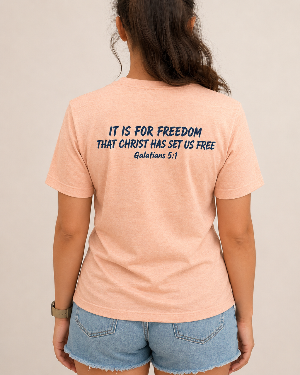 Freedom Galatians 5:1 Bella+Canvas Heather Peach tee with BLOVD Heritage Navy Scripture artwork, approved female warm-neutral studio back view showing the back of the neck with denim shorts visible.